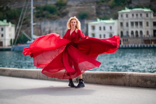 A woman in a red dress, a fashion model with long silk wings in a flowing dress, flying fabric on the embankment