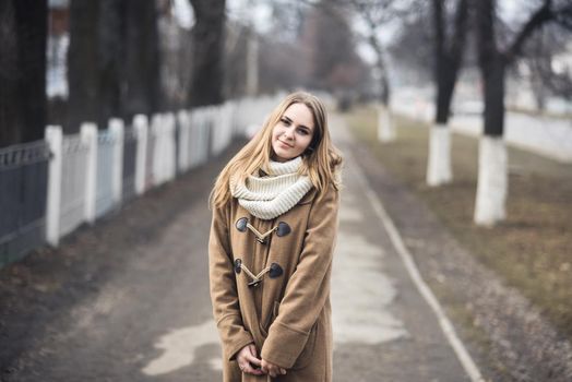 Blond-haired young girl cheerful joyful dressed you in a beige coat and white scarf. Autumn gray street on the background. empty road