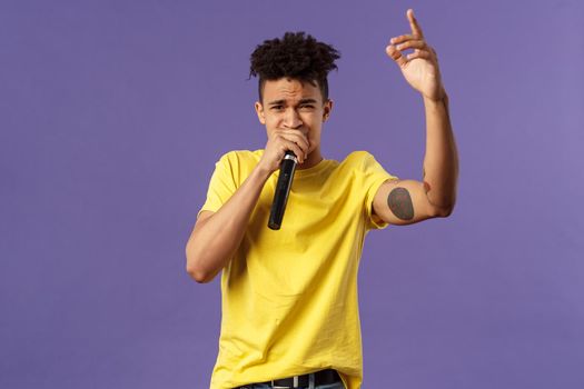 Portrait of young carefree sassy hipster guy with tattoos, yellow t-shirt performing in front audience with his hip-hop or rap, singing song, raising finger up, hold microphone, karaoke.