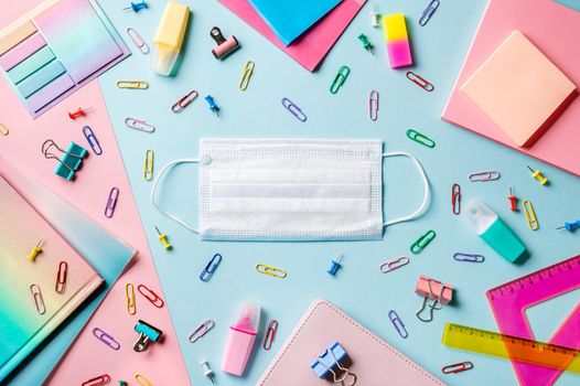 Protective medical mask on creative student desk with colorful modern school supplies on blue and pink duotone background. Top view. Flat lay. Copy space. Back to school covid19 concept