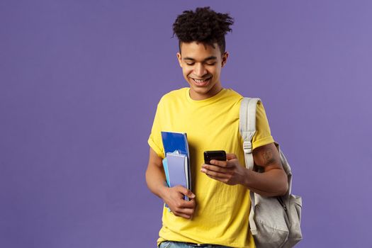 Back to school, university concept. Portrait of pleased good-looking male student texting friend, looking at mobile phone on his way to classes in campus, hold notebooks and backpack.