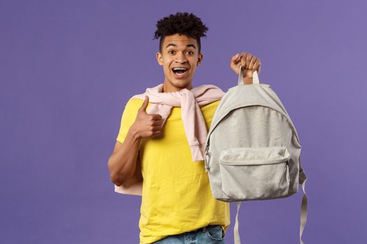 Portrait of cheerful young handsome male student, man recommending backpack, holding rucksack and show thumbs-up, bought new equipment for university semester, purple background.