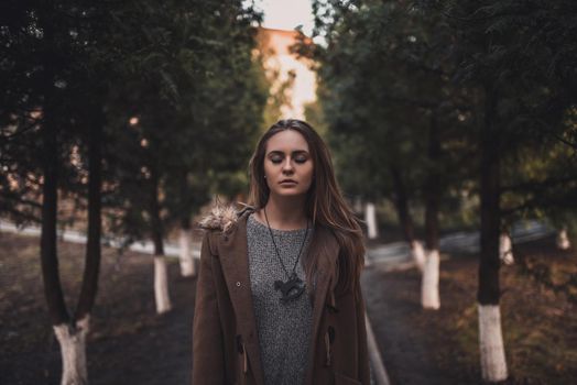 beautiful young blonde model girl. White pants. gray knitted sweater. black boots. . wooden pendant on the neck in the form of a horse. in a brown coat posing. On the Sunset. Portrait. near the trees