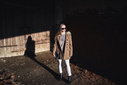 beautiful young blonde model girl. White pants. gray knitted sweater. black boots. black sunglasses. wooden pendant on the neck in the form of a horse. in a brown coat posing. On the Sunset. Portrait