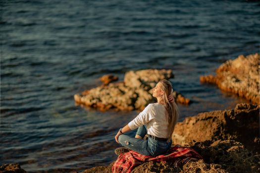 Attractive blonde Caucasian woman enjoying time on the beach at sunset, sitting in a blanket and looking to the side, with the sunset sky and sea in the background. Beach vacation