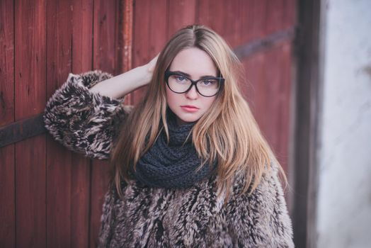 Girl blonde in cat eyes glasses in an artificial faux fur coat posing. Red orange pink tones in the photo. Around the old shabby abandoned buildings. Cold winter. wooden wall