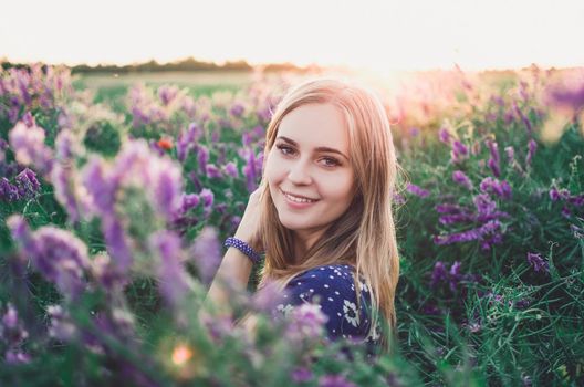 Light-skinned happy smiling European blonde young slim girl walks on tall grass overgrown with purple flowers. Women's blue short skin-tight dress with white daisy print.
