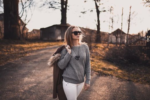 beautiful young blonde model girl. White pants. gray knitted sweater. black boots. black sunglasses. wooden pendant on the neck in the form of a horse. in a brown coat posing. On the Sunset. Portrait