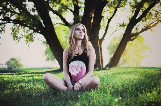 Lovely young blonde girl with long curly hair on a background of Tall Green Tree. A woman is wearing a cropped black top with a white print and pink lettering and gray short high-waisted shorts.