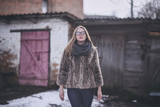 Girl blonde in cat eyes glasses in an artificial faux fur coat posing. Lying snow on the background. Red orange pink tones in the photo. Around the old shabby abandoned buildings. Cold winter