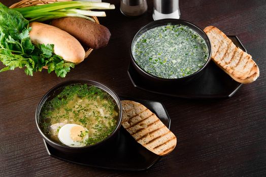 Okroshka, broth with noodles and eggs in black plate, toasted bread, herbs on dark wooden table. Healthy seasonal lunch