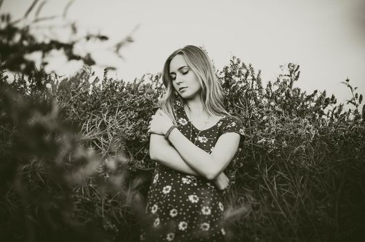 A young blonde European thin girl stands in the midst of a very tall purple and poppy plant hugs herself with her eyes closed. Women's blue short skin-tight dress with white daisy print.