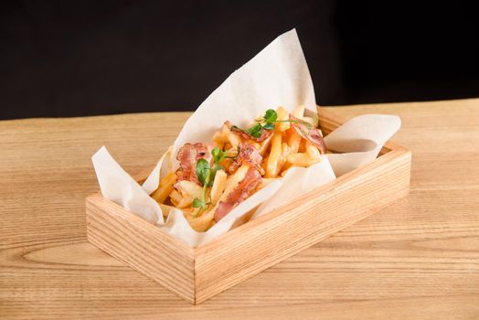 French fries with fried bacon on parchment in wooden box on the table.