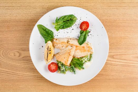 Grilled chicken fillet with basil, cherry tomatoes, lemon, peas, sauce on white round plate on wooden table.