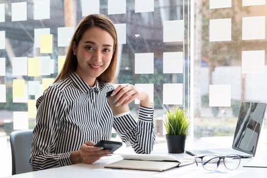 Beautiful young Asian businesswoman drinking a coffee and holding a smartphone working on laptop at office. Looking at camera..