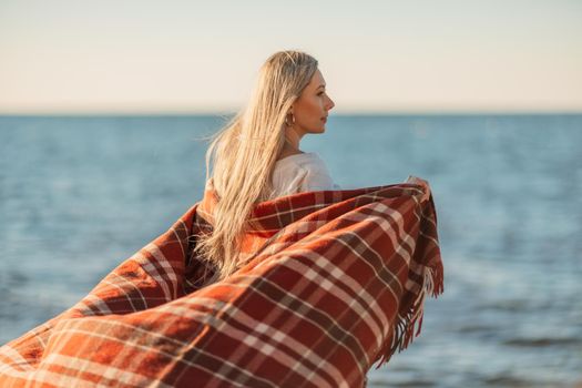 Attractive blonde Caucasian woman enjoying time on the beach at sunset, walking in a blanket and looking to the side, with the sunset sky and sea in the background. Beach vacation.