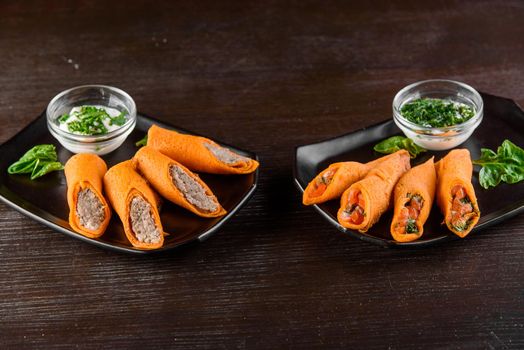 Orange pancakes with pate, salmon, dill, sour cream on black plate on a wooden table. Filled rolls.