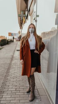 young ukrainian blonde woman in protective medical mask kn95 respirator on street against background of shop windows. coronavirus infection. accessory covid-19