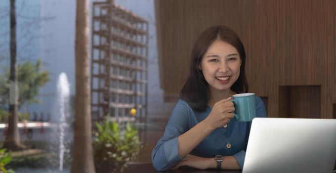Smiling Asian businesswoman holding a coffee mug and laptop at the office. Looking at the camera..