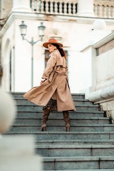 Outdoor fashion portrait of young elegant fashionable brunette woman, model in stylish hat, choker and light raincoat posing at sunset in European city