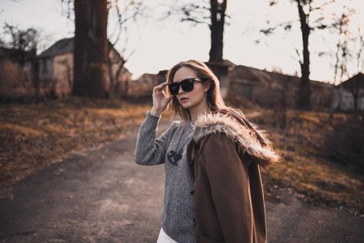 beautiful young blonde model girl. White pants. gray knitted sweater. black boots. black sunglasses. wooden pendant on the neck in the form of a horse. in a brown coat posing. On the Sunset. Portrait