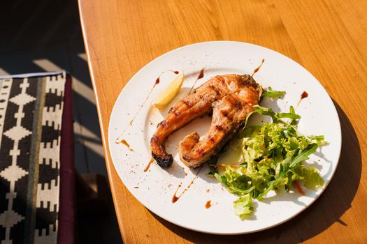 Grilled salmon steak with lettuce and lemon on a white plate on a wooden table