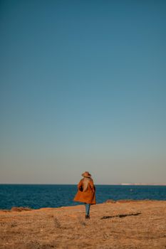 A woman walking along the coast near the sea. An elegant lady in a brown coat and a hat with fashionable makeup walks on the seashore.