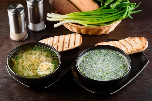 Okroshka, broth with noodles and eggs in black plate, toasted bread, herbs on dark wooden table. Healthy seasonal lunch