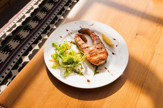 Grilled salmon steak with lettuce and lemon on a white plate on a wooden table