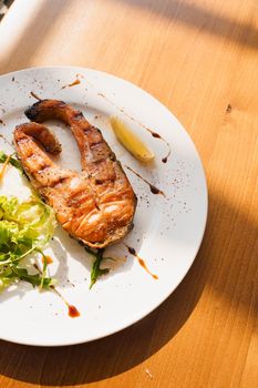 Grilled salmon steak with lettuce and lemon on a white plate on a wooden table