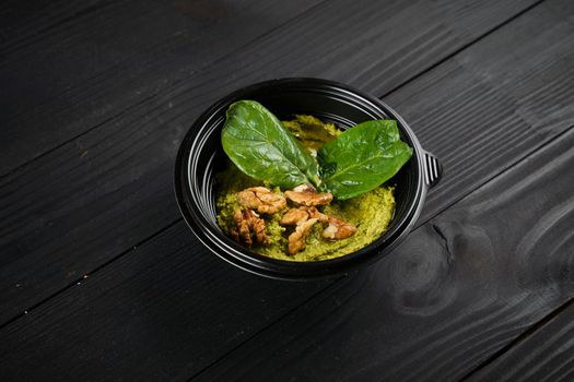 Hummus with basil and walnuts garnished with herbs in black bowl on dark wooden table.