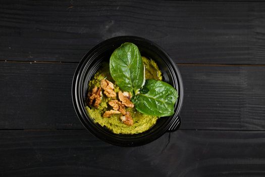 Hummus with basil and walnuts garnished with herbs in black bowl on dark wooden table.
