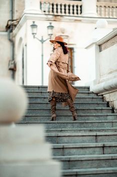 Outdoor fashion portrait of young elegant fashionable brunette woman, model in stylish hat, choker and light raincoat posing at sunset in European city