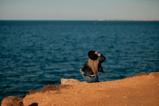 A serious blonde girl in a stylish black leather jacket and a black hat is sitting with her back to the seashore.