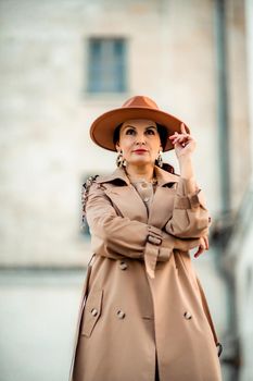 Outdoor fashion portrait of young elegant fashionable brunette woman, model in stylish hat, choker and light raincoat posing at sunset in European city