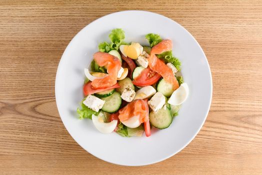 Salad with salmon, feta cheese, egg, tomato, cucumber, lettuce on white plate on light wooden table.