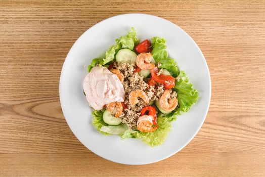 Salad with shrimps, quinoa, tomatoes, peppers, cucumber, lettuce, mayonnaise on white round plate on wooden table.