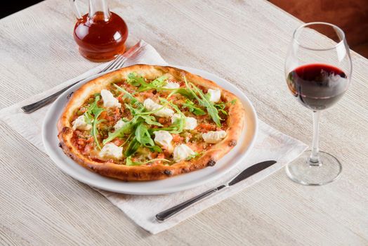 Pizza with salmon, feta cheese, sauce, tomatoes, on white round plate on light wooden table on background of glass of red wine.