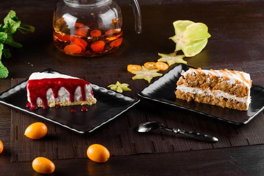 Cheesecake with berry sauce and carrot cake with sour cream and caramel on black square plates on dark wooden table.