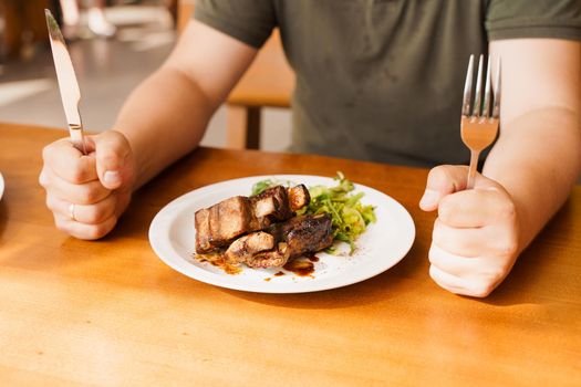 Grill ribs with salad on a white plate on a wooden table. Hungry visitor holding a fork and knife.