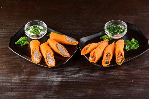 Orange pancakes with pate, salmon, dill, sour cream on black plate on a wooden table. Filled rolls.