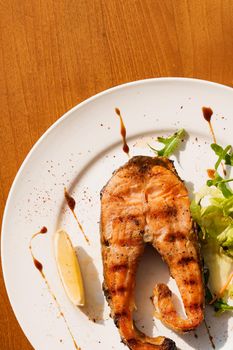 Grilled salmon steak with lettuce and lemon on a white plate on a wooden table