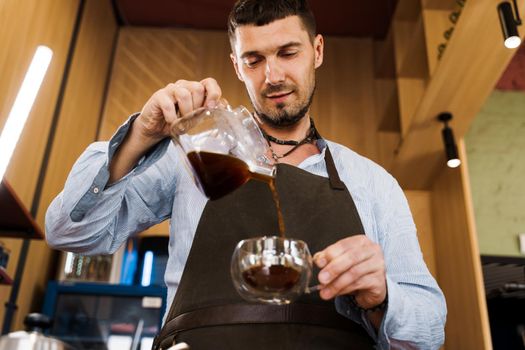 Handsome barista pours flavored coffee in glass pot in cafe. Coffee brewing syphon and aeropress alternative methods