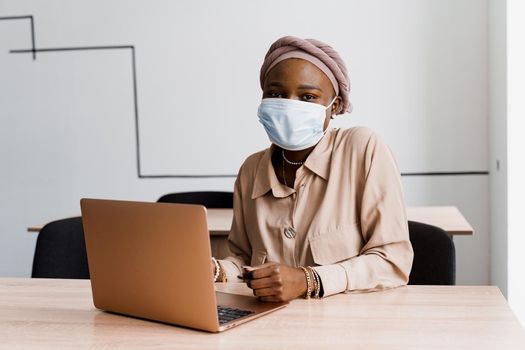 African black woman with laptop. Using computer for on-line work. Medical mask for protection against coronovirus covid-19.