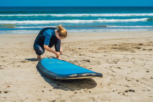 Summer time and active rest concept. Young surfer woman beginner fastens leash across leg, going to surf on big barral waves on ocean, dresseed in boardies, poses aganist sky on hot sunny day.