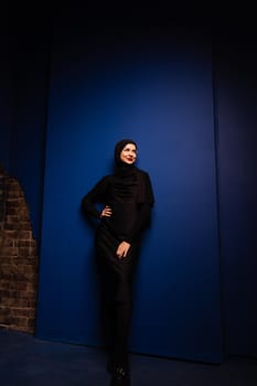 Fashion muslim woman in black hijab is posing on blue background in studio. Professional caucasian model