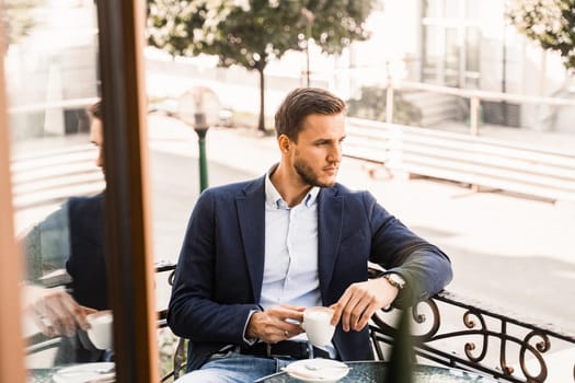 Man is sitting on the summer terrace in cafe, drinking coffee and smiling. Handsome man with cup of coffee in cafe. Morning lifestyle of male