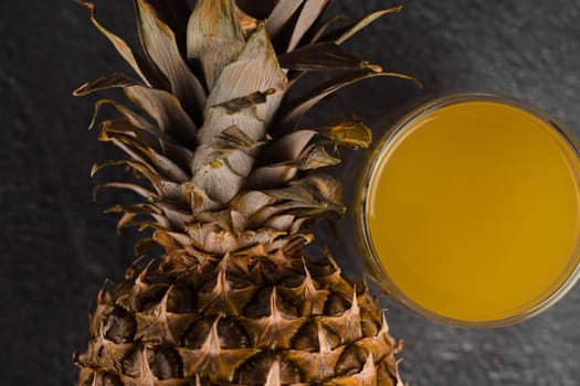 Pineapple fruit and juice in double glass cup on black stone background. Pouring yellow tropical fruit juice into glass.