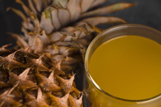 Pineapple fruit and juice in double glass cup on black stone background. Pouring yellow tropical fruit juice into glass.