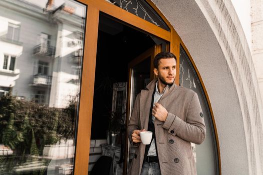 Handsome man with cup of coffee posing in coat in cafe. Male lifestyle